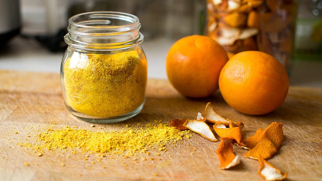 Glass jar filled with yellow powder next to whole oranges and dried orange peel on a wooden surface, warm citrus tones