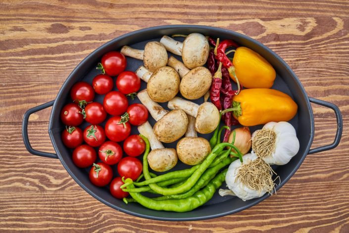 An oval dark pan filled with cherry tomatoes, mushrooms, chili peppers, yellow bell peppers and garlic bulbs on a wooden surface